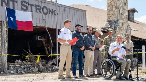 حاكم تكساس جريج أبوت يتحدث في مؤتمر صحفي بعد الفيضانات، محاطًا بمسؤولين محليين، مع العلم التكساسي خلفه.