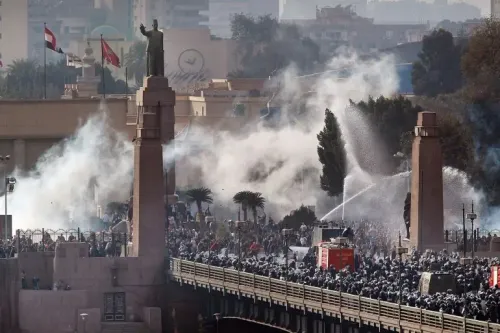 تجمع حشود كبيرة من المتظاهرين في وسط القاهرة، وسط دخان الغاز المسيل للدموع، مع وجود تمثال بارز في الخلفية.