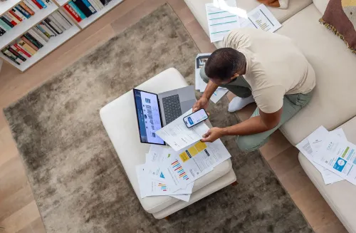 رجل يجلس على أريكة محاطة بأوراق مالية وجهاز كمبيوتر محمول، يتفقد بيانات استثمارية على هاتفه، يعكس مفهوم استراتيجية KISS للاستثمار.