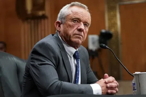 رجل مسن يجلس في قاعة جلسات، يبدو عليه التفكير العميق، يرتدي بدلة رسمية، مع وجود ميكروفون أمامه، في سياق مناقشة حول اللقاحات.