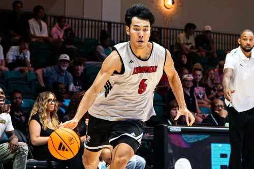 لاعب كرة السلة الصيني Zhang Boyuan يرتدي قميص Louisville Cardinals ويحمل الكرة خلال مباراة في NCAA، مع جمهور ومشجعين في الخلفية.