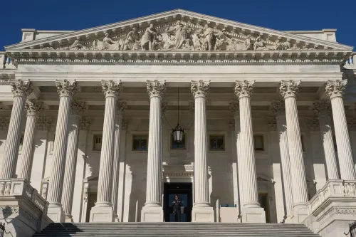 واجهة مبنى الكابيتول الأمريكي، تظهر الأعمدة الرخامية والنقوش المعمارية، مع وجود حارس عند المدخل، تعكس أهمية الحكومة الأمريكية.