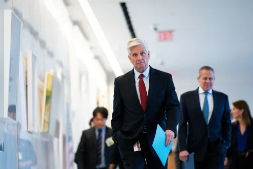 رجل يرتدي بدلة رسمية يسير في ممر مع مجموعة من الأشخاص، مع وجود لوحات فنية على الجدران، مما يعكس أجواء مهنية.