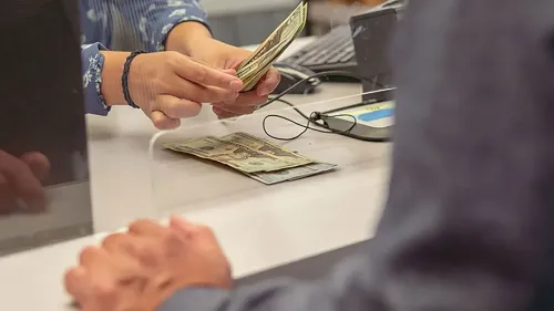 شخص يقوم بتسليم النقود من خلال مكتب مصرفي، مع التركيز على أهمية إدارة المدخرات والعوائد المالية.