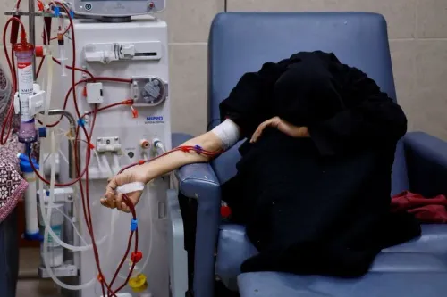 امرأة ترتدي ملابس سوداء تجلس على كرسي في مستشفى، تتلقى العلاج الكيميائي، بينما يظهر جهاز طبي بجانبها. تعكس الصورة معاناة الأمهات في غزة.