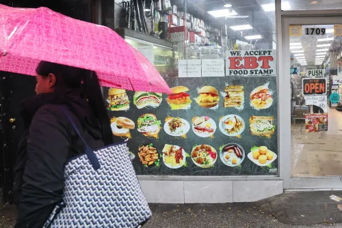 امرأة تحمل مظلة وردية تسير بجوار واجهة متجر تعرض لافتة "نقبل بطاقات الطعام EBT" وصور أطباق متنوعة، تعبيرًا عن تأثير الإغلاق الحكومي على الإعانات الغذائية.