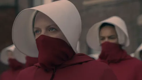 امرأة ترتدي رداءً أحمر وقبعة بيضاء، تمثل شخصية "الخادمة" من مسلسل "The Handmaid's Tale"، مع خلفية لمجموعة من النساء الأخريات.