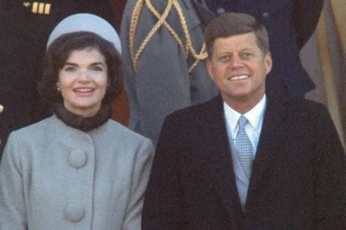 جاكلين كينيدي ترتدي معطفًا رماديًا وقبعة أنيقة أثناء حفل تنصيب زوجها جون كينيدي، مبتسمة بجانب الرئيس. تعكس إطلالتها أسلوبًا عصريًا وجاذبية.