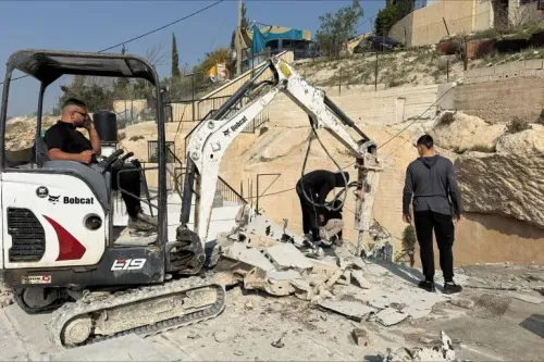 عمال يستخدمون آلة هدم صغيرة لتفكيك أجزاء من منزل في صور باهر، بينما يظهر شخص آخر يراقب العملية، مع خلفية منازل في القدس الشرقية.