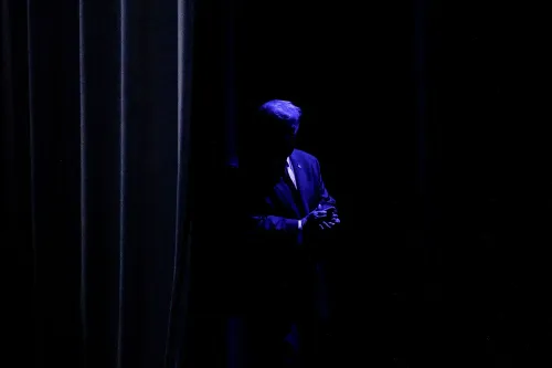 شخصية سياسية بارزة تقف في الظل خلف ستارة، مما يعكس التوترات السياسية الحالية وتأثيرها على المجتمع الأمريكي.