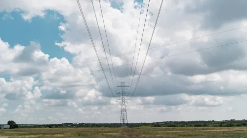 أبراج كهرباء عالية تمر عبر حقل مفتوح تحت سماء ملبدة بالغيوم، تعكس التحديات في تحديث البنية التحتية الكهربائية وارتفاع تكاليف الطاقة.