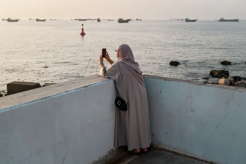 امرأة ترتدي الحجاب تقف على حافة رصيف تطل على البحر، تلتقط صورة بهاتفها، بينما تظهر سفن في الخلفية.