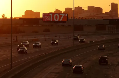 غروب الشمس يضيء مدينة مزدحمة، حيث تسير السيارات على الطريق السريع، مع عرض درجة الحرارة 107°F. تعكس الصورة التحديات المناخية التي تواجهها الولايات المتحدة.