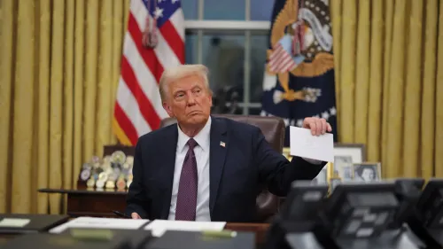 الرئيس دونالد ترامب يجلس خلف مكتبه في المكتب البيضاوي، ممسكًا بورقة بيضاء، بينما تلوح خلفه الأعلام الأمريكية.