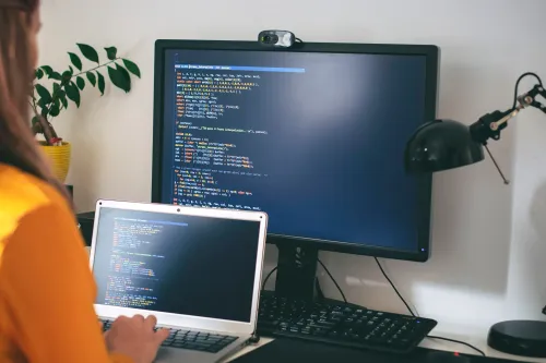 مهندسة برمجيات تكتب شيفرة برمجية على حاسوب محمول وشاشة كبيرة، تعبيراً عن تحديات استخدام أدوات الذكاء الاصطناعي في تطوير البرمجيات.