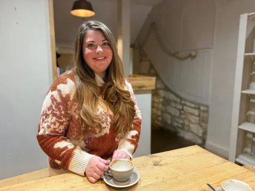 امرأة شابة تجلس في مقهى ريفي بكوتسوولدز، ترتدي سترة ملونة وتمسك بفنجان قهوة، تعكس أجواء الحياة الهادئة في المنطقة.