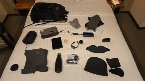 تظهر الصورة مجموعة من الأغراض الشخصية المعبأة على سرير، تشمل حقيبة ظهر، ملابس، زجاجة ماء، وأدوات تقنية، تشير إلى استعدادات السفر.