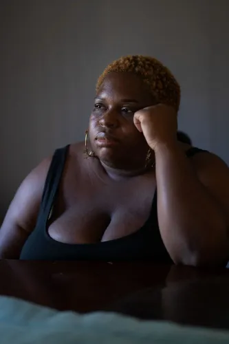 امرأة ذات بشرة داكنة تجلس في غرفة مضاءة بشكل خافت، تعبر عن مشاعر القلق والتفكير العميق، مما يعكس التوتر المرتبط بالقرارات الطبية أثناء الحمل.