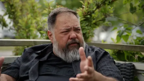 رجل ذو لحية رمادية يتحدث بجدية في حديقة، مع خلفية من النباتات الخضراء، يعبر عن أفكاره حول الذكاء الاصطناعي وتأثيراته.