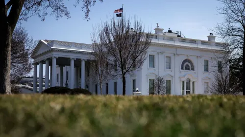 واجهة البيت الأبيض مع العلم الأمريكي يرفرف فوقه، محاطة بأشجار جرداء وحديقة خضراء، تعكس أجواء السياسة الأمريكية.