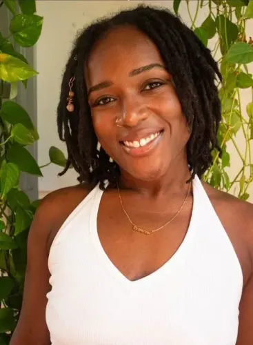 امرأة مبتسمة تحمل شعرًا مجعدًا، ترتدي قميصًا أبيض، وتقف أمام حائط مزين بالنباتات. تعكس الصورة الأمل والإيجابية في سياق الاحتجاجات ضد قوانين الهجرة.