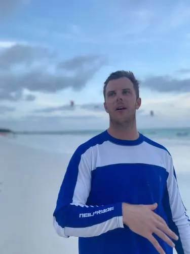 لوكاس هانتر، شاب أمريكي يرتدي قميصاً أزرق، يتحدث على شاطئ في كولومبيا، حيث كان يخطط لرحلة لتجربة ركوب الأمواج الشراعية.