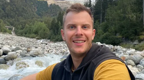 لوكاس هانتر مبتسم بجانب مجرى مائي في منطقة جبلية، محاط بالأشجار والصخور، يعكس حبه للمغامرة والطبيعة.
