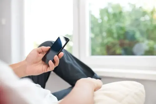 شخص يجلس على أريكة ويتصفح هاتفه المحمول بجوار نافذة تطل على منظر طبيعي، مما يعكس أجواء الاسترخاء قبل النوم.