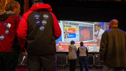 مجموعة من الأشخاص يشاهدون شاشة كبيرة تعرض نتائج الانتخابات الأمريكية، مع تفاصيل عن ولايات مثل ويسكونسن وأوهايو.