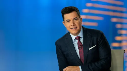 توم لالماس، المذيع الجديد لبرنامج "أخبار إن بي سي نايتلي نيوز"، مبتسمًا في استوديو الأخبار، خلفية زرقاء مضيئة.