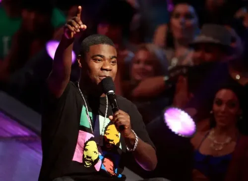 تريسي مورغان يتحدث على المسرح مرتديًا تيشيرت ملون، بينما يستمتع الجمهور خلفه بعرضه الكوميدي.