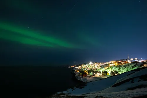 منظر ليلي لقرية في غرينلاند، مضاءة بأضواء دافئة تحت سماء مليئة بالنجوم، مع شريط من الأضواء الشمالية الخضراء في الخلفية.