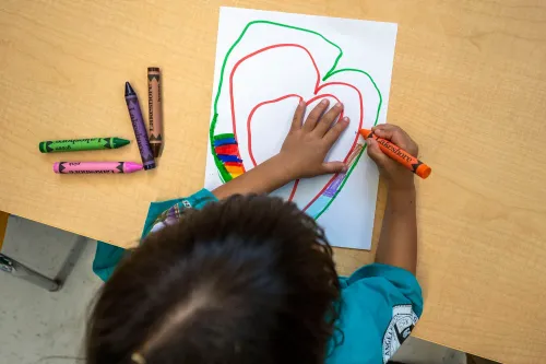 طفل يستخدم أقلام تلوين لرسم قلب على ورقة، مع وجود أقلام ملونة أخرى حوله، مما يعكس أهمية الفنون في رعاية الأطفال.