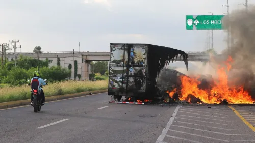 شاحنة محترقة على جانب الطريق، بينما يراقب راكب دراجة نارية الحادث، مما يعكس تصاعد العنف من عصابات المخدرات في المكسيك.