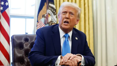 رجل يرتدي بدلة زرقاء وقميص أبيض مع ربطة عنق زرقاء، يتحدث بجدية أثناء جلوسه في مكتب رسمي، مع العلم الأمريكي خلفه.