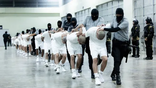 رجال يرتدون زيًا أبيض، مُقيدون ويُقادون من قبل أفراد أمن في مرفق احتجاز، في سياق عمليات الترحيل للمهاجرين.