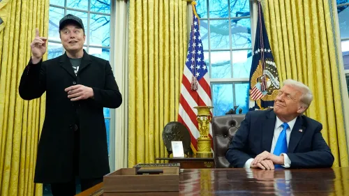 إيلون ماسك يتحدث في المكتب البيضاوي بينما ينظر دونالد ترامب إليه بابتسامة، مع العلم الأمريكي خلفهم.