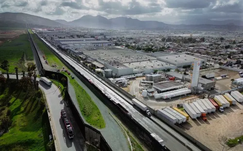 صورة تظهر الحدود بين الولايات المتحدة والمكسيك، مع شاحنات تجارية تنتظر العبور، تعكس العلاقات التجارية المتوترة بين البلدين.