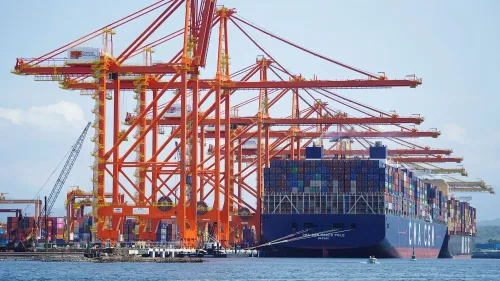 سفينة حاويات كبيرة تخضع للتفريغ في ميناء، مع رافعات حمراء ضخمة وآلاف الحاويات المتنوعة، تعكس التبادل التجاري بين أمريكا وكندا والمكسيك.