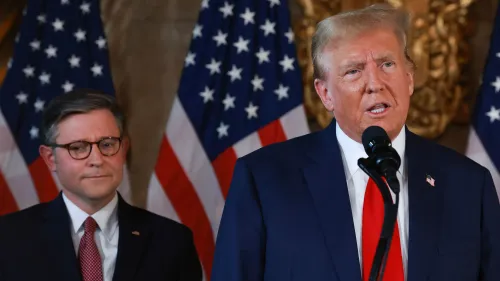 ترامب يتحدث في مؤتمر صحفي حول إمكانية زيادة الضرائب على الأثرياء، بينما يقف مستشار خلفه، مع أعلام أمريكية في الخلفية.