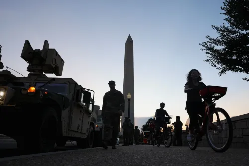 جنود يرتدون زيًا عسكريًا يقفون بجانب مركبات عسكرية، بينما يمر راكبون على دراجات في واشنطن، مع وجود نصب واشنطن في الخلفية.
