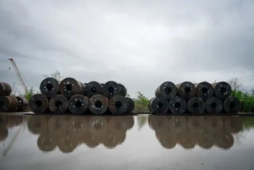 صورة لأسطوانات معدنية متراصة بجانب بركة مياه تعكس السحب الرمادية في السماء، تعبر عن تأثير التعريفات الجمركية على الصناعة.