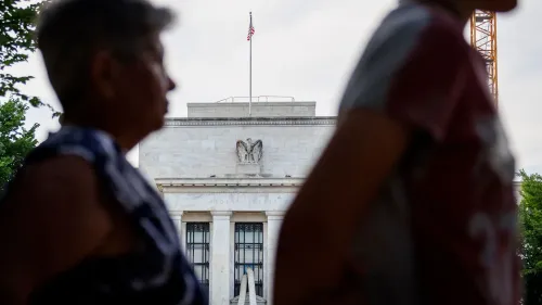 شخصان يسيران أمام مبنى الاحتياطي الفيدرالي الأمريكي، مع العلم الأمريكي يرفرف في الخلفية، في سياق مناقشات حول السياسة النقدية والتضخم.