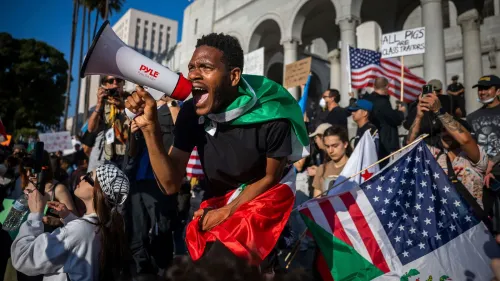 متظاهر يحمل مكبر صوت ويصرخ في حشد خلال احتجاج في لوس أنجلوس، مع أعلام متعددة في الخلفية، تعبيرًا عن الغضب والمشاركة.