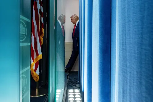 صورة تُظهر دونالد ترامب واقفًا في ممر ضيق خلف ستائر زرقاء، مع العلم الأمريكي في الجهة اليسرى، مما يرمز إلى التوترات السياسية والاقتصادية الحالية.