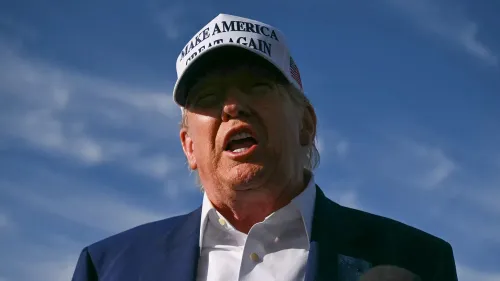 ترامب يتحدث بحدة في مناسبة يوم الذكرى، مرتديًا قبعة تحمل شعار "اجعلوا أمريكا عظيمة مرة أخرى"، مع سماء زرقاء في الخلفية.