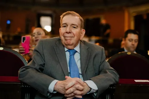 رجل في بدلة رمادية يجلس مبتسمًا في قاعة، مع وجود مشاهدين آخرين في الخلفية. الصورة تعكس أجواء سياسية في سياق الأحداث الفنزويلية.