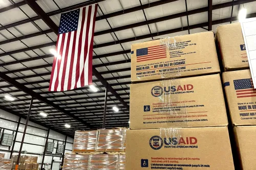 صناديق مساعدات تحمل شعار الوكالة الأمريكية للتنمية الدولية (USAID) في مستودع، مع العلم الأمريكي يتدلى في الخلفية.