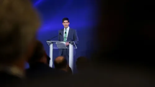 كيفن وارش يتحدث في مؤتمر، مع وجود جمهور خلفه. يبرز في الخلفية ضوء أزرق، مما يضيف جوًا احترافيًا للحدث.