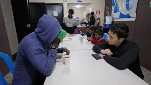 رجل يرتدي سترة زرقاء وقبعة خضراء يجلس مع شخص آخر في مركز للاجئين في غواتيمالا، محاطون بمهاجرين آخرين. تعبيرات الوجوه تعكس القلق والوضع الصعب.